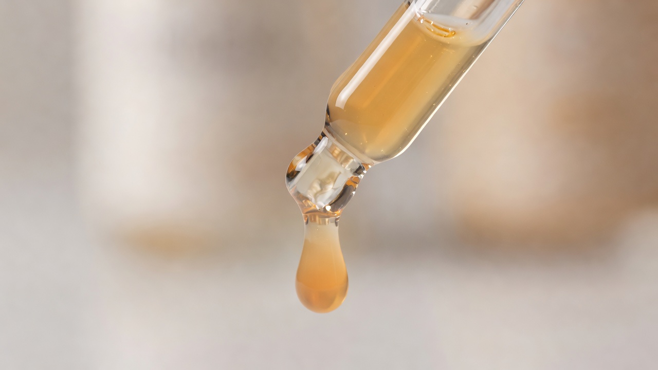 Macro shot of a glass dropper tip releasing a pale orange vitamin C serum drop against a blurred background